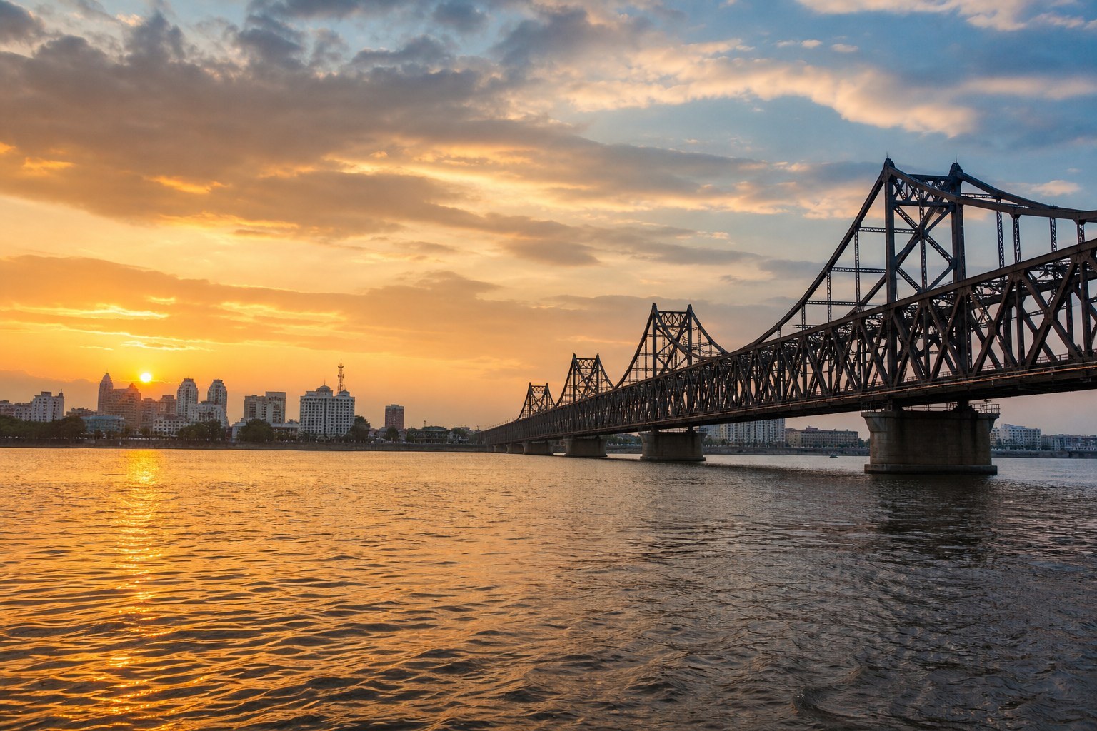 Water-level sunset view toward the historic Yalu River bridge and Dandong skyline
