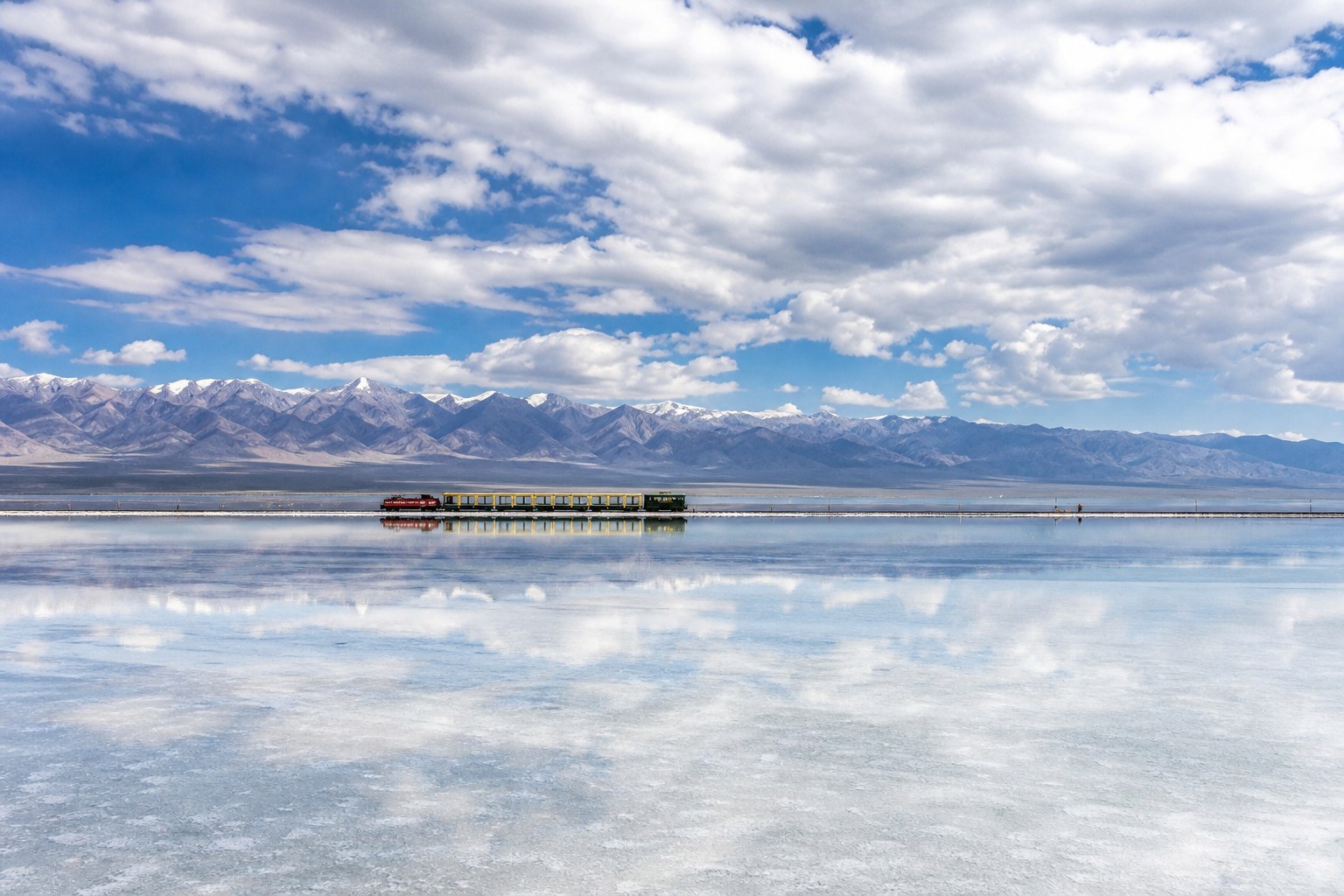 Small scenic train crossing reflective Chaka Salt Lake beneath a mirror sky